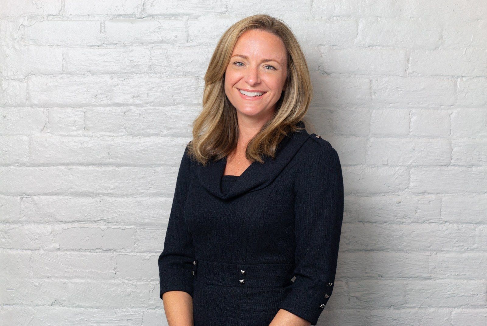 Amy Clark, founder of Amy K Clark Design, in a navy dress against a white-painted brick wall