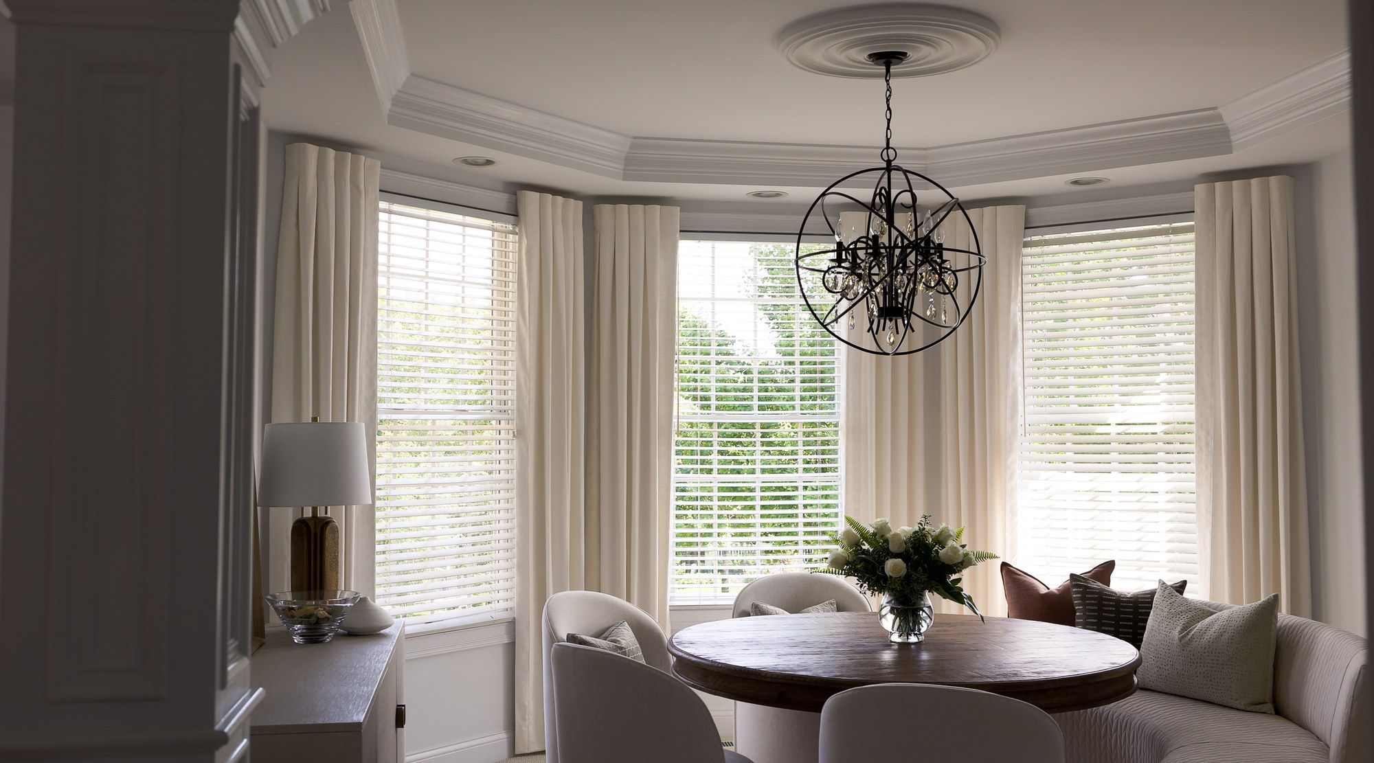 Ivory pinch-pleat drapery and white wood blinds dressing three bay windows in a breakfast room, beneath a wire-orb chandelier and ceiling medallion