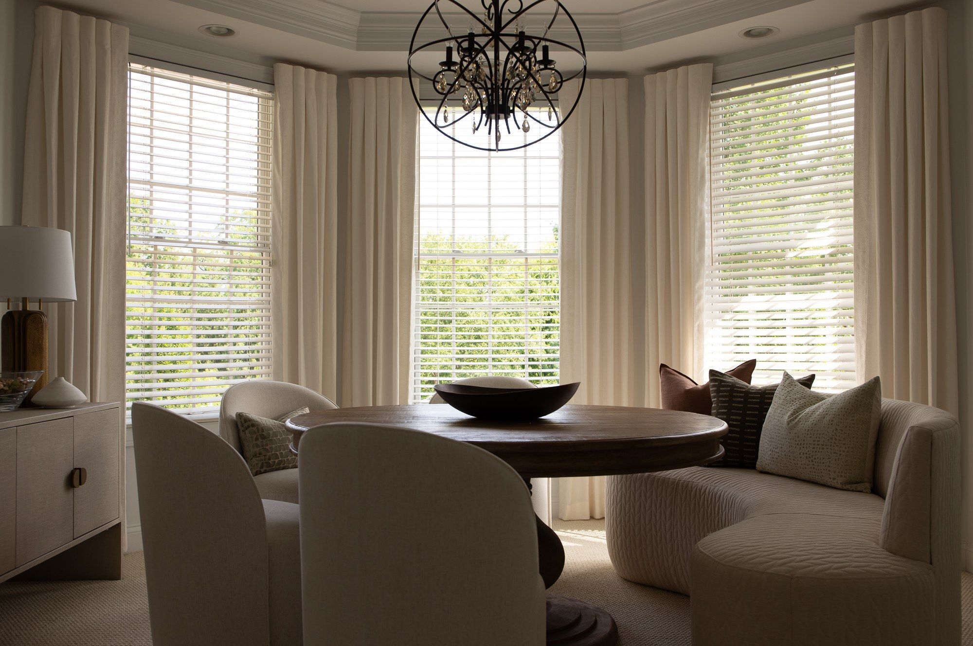 Ivory pinch-pleat drapery and white wood blinds dressing bay windows in a breakfast room, with dining chairs set beneath a wire-orb chandelier