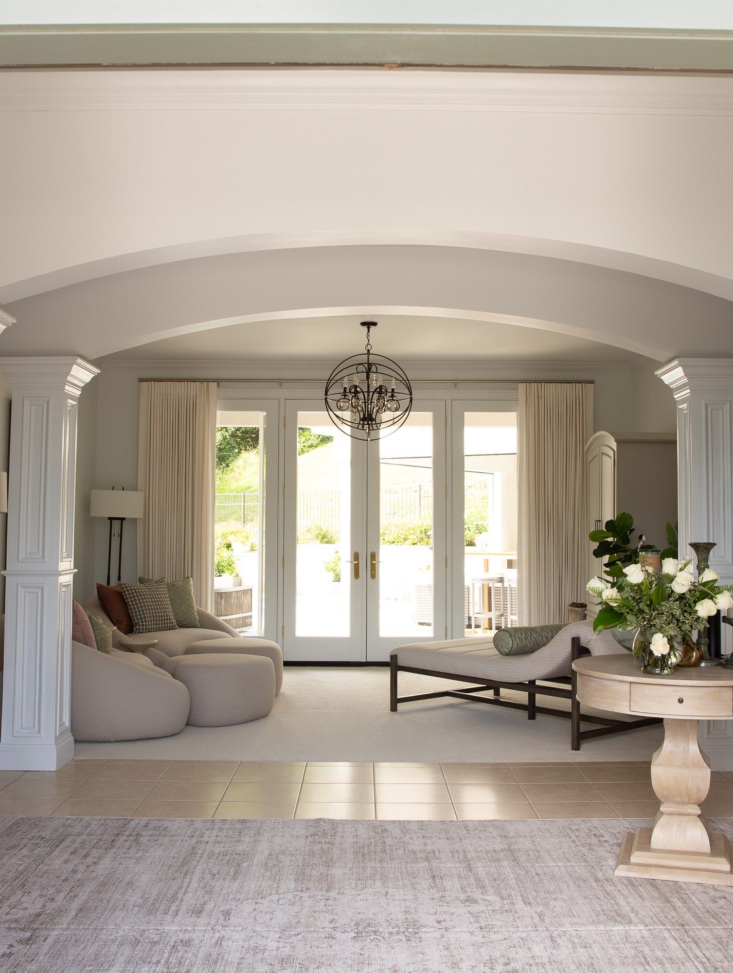 A living room seen through a columned archway, ivory pinch-pleat drapery framing French doors beneath a wire-orb chandelier