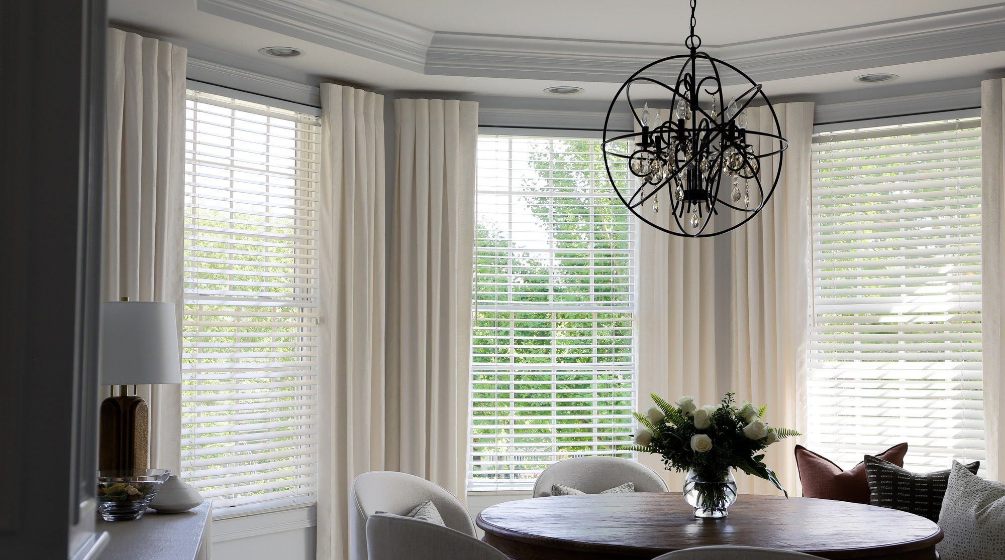 White wood blinds beneath ivory pinch-pleat drapery in a bay-window breakfast room, with a wire-orb chandelier overhead